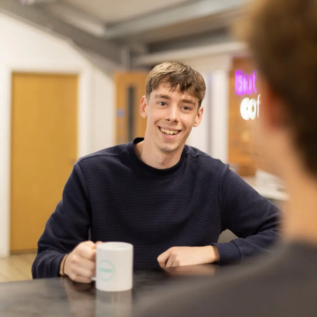 An image of Charles smiling while talking to someone at a desk with a PMW mug.