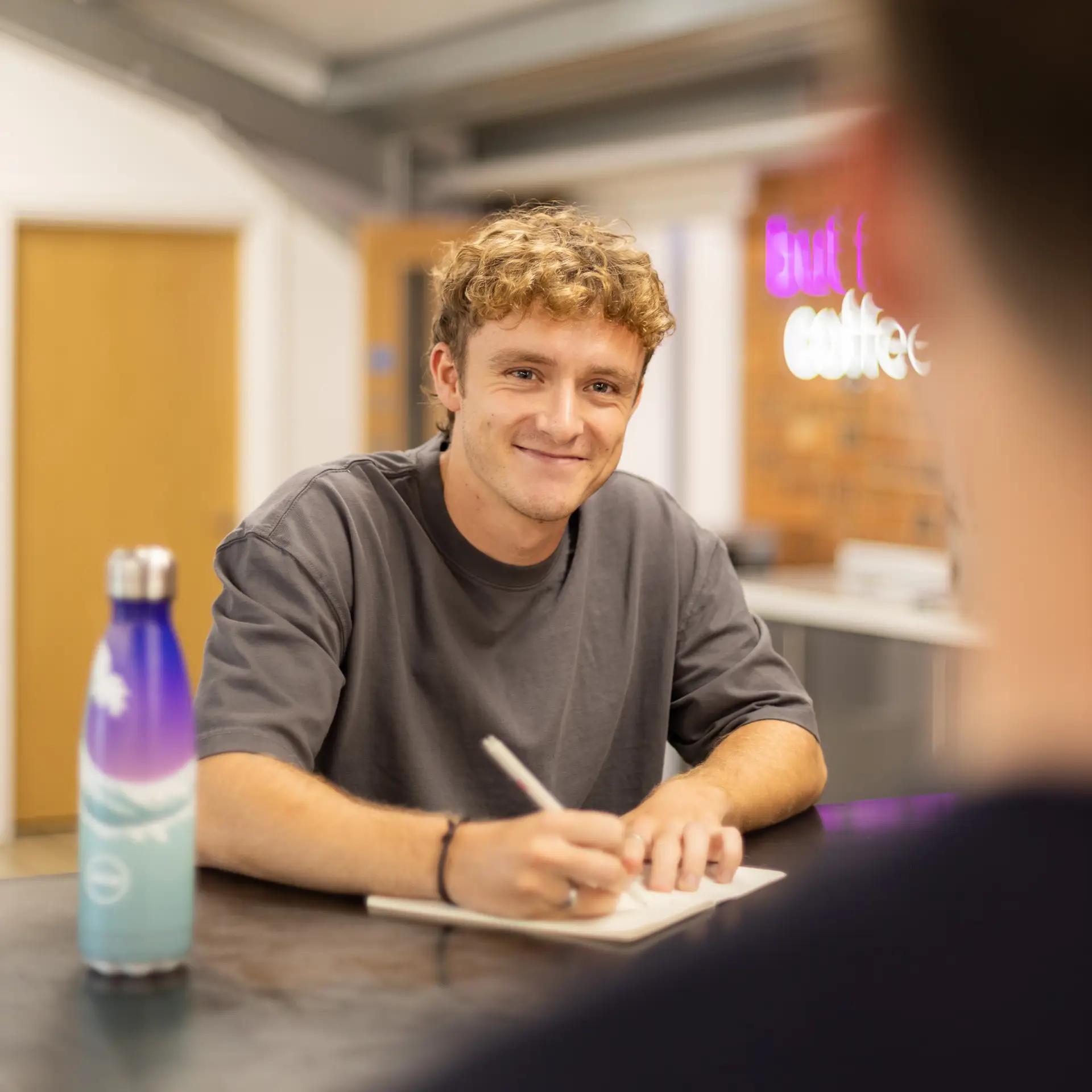 A picture of Charlie sitting writing at a table with a water bottle talking to a colleague who is out of view.