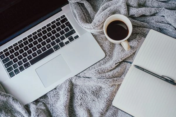 A view from above -A laptop on a cozy grey blanket with a cup of black coffee from above and an open notebook.