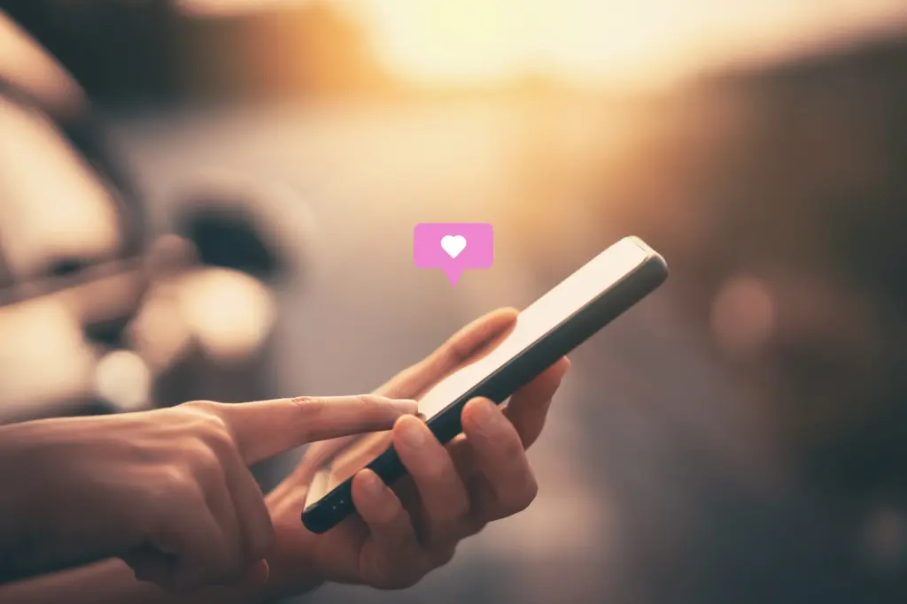 Close up of hand scrolling on a phone with a love heart reaction in the style of a speech bubble emerging from the top. Warm, sunset tones.